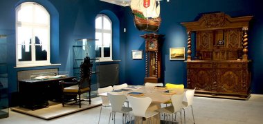 Das Danzig Zimmer Ein Raum mit blauen Tapeten und alten Möbelstücken dekoriert. Von der Decke hängt ein beleuchtetes Segelschiff