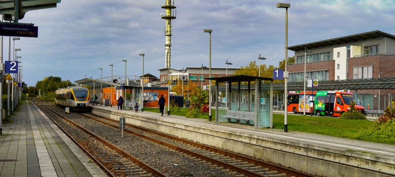 Hier ist der Warendorfer Bahnhof mit einem hereinfahrenden Zug sowie Bussen im Hintergrund abgebildet.