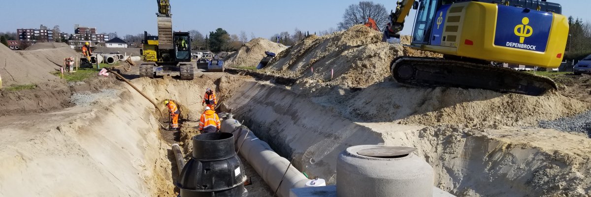 Kanalbauarbeiten In de Brinke Eine geböschte Baugrube mit einer Tiefe von ca. 2 Meter in der einige Arbeiter zwei Leitungen verlegen. Im Vordergrund sind zwei Schachtbauwerke errichtet worden.