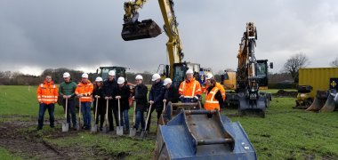 Erster Spatenstich In de Brinke Aufstellung von Menschen und Maschinen für den ersten Spatenstich. Die Vertreter von verschiedenen Projektbeteiligten tragen Helme und haben Spaten in der Hand.